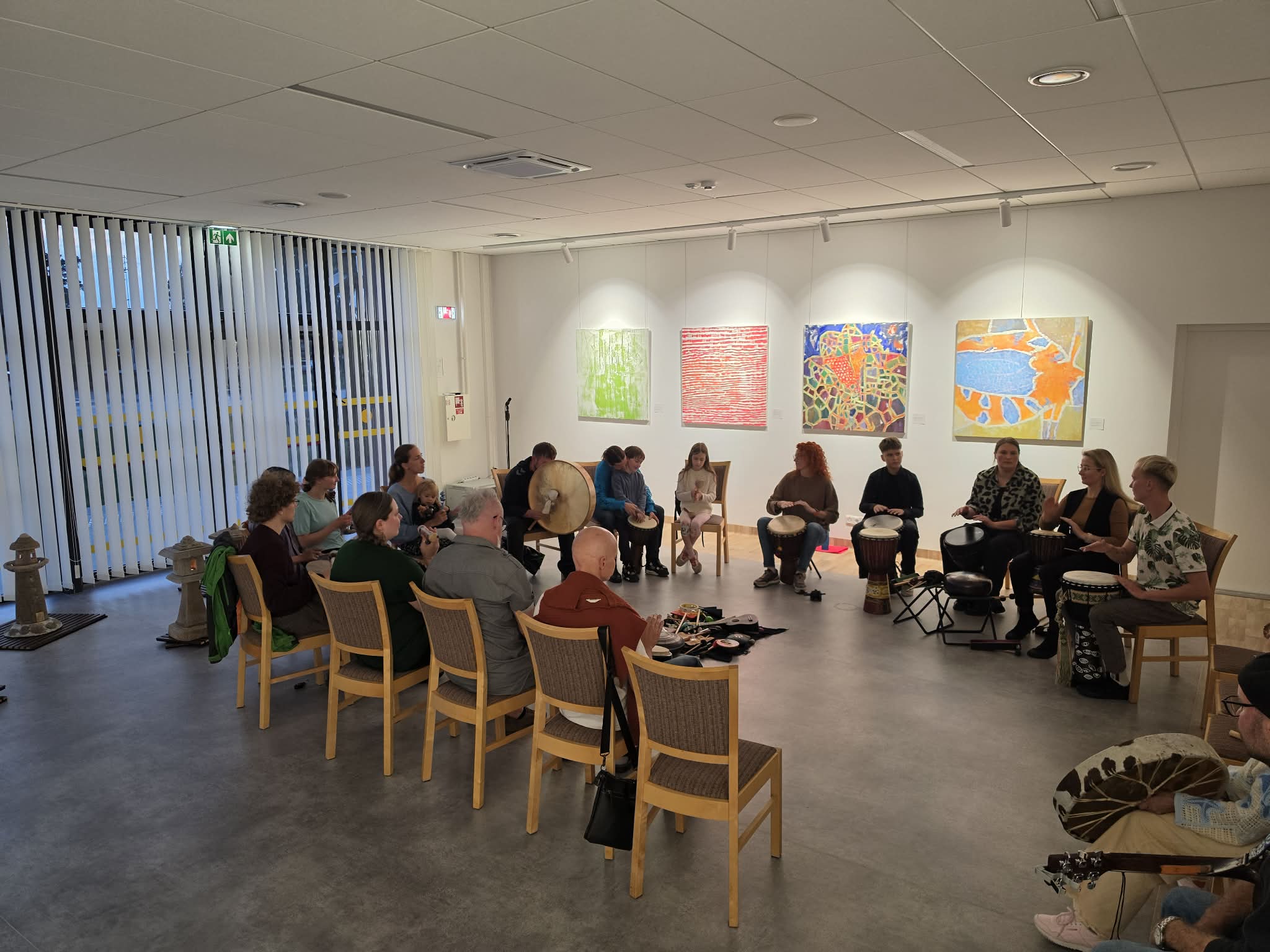 Adults and children play percussion in a circle, with four paintings behind them